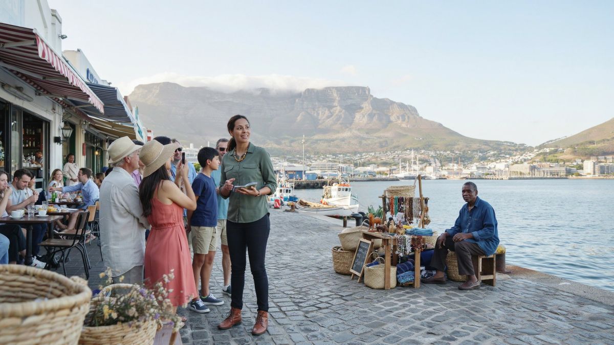 South africa tourism - travel and tour world A tour guide welcomes visitors beside a craft stall at cape town’s v&a waterfront, with table mountain rising behind the harbour.