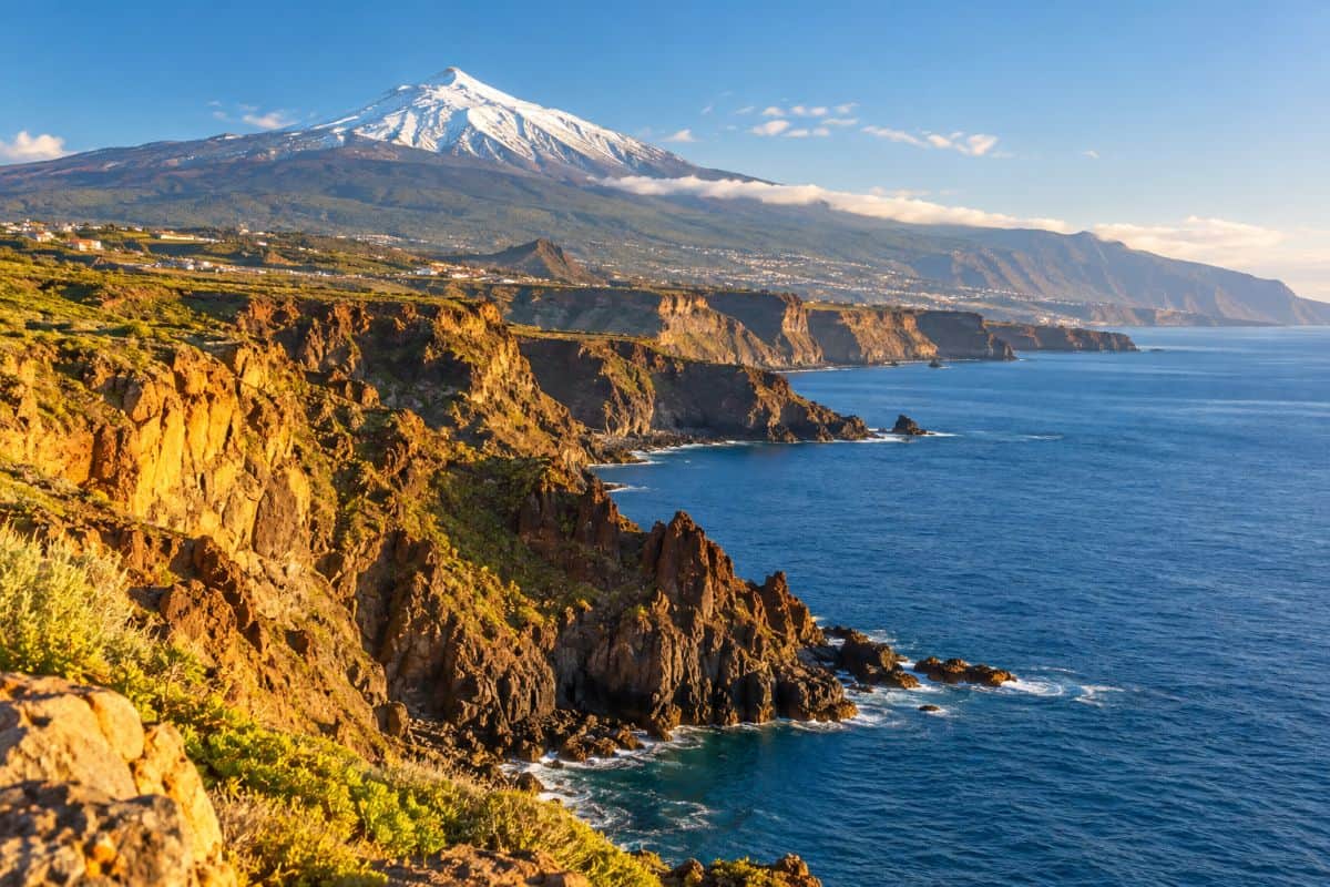Spain and greece are great for winter travel with sun in the canaries and mountain adventures in crete - travel and tour world Sunlit volcanic cliffs along tenerife’s blue atlantic coast, with snow-capped mount teide rising in the background.