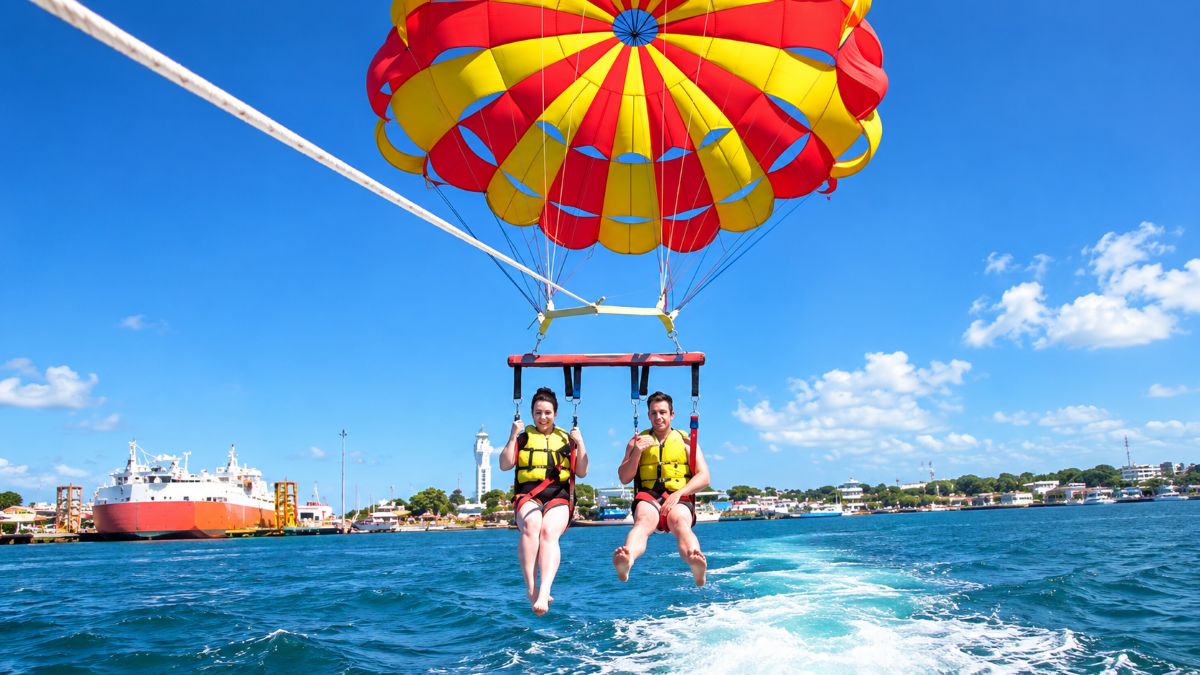 Two tourists parasailing over bright blue ocean water beneath a colourful red and yellow canopy, with a coastal harbour and ships in the background.