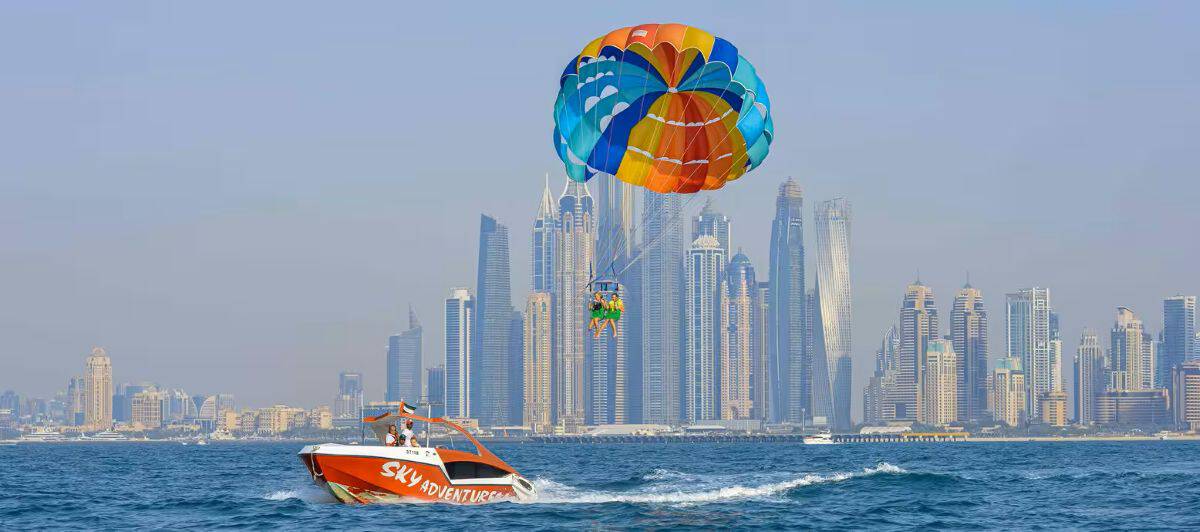 Two people parasailing beneath a colourful canopy above the sea, with a speedboat below and dubai&rsquo;s skyline in the background.