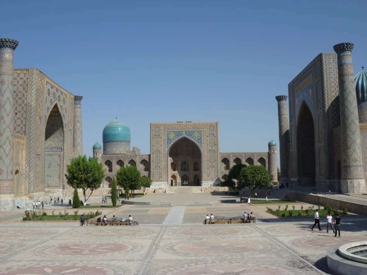 A wide-angle view of the registan square in samarkand, uzbekistan, showcasing grand madrasahs with intricate tile work, towering minarets, and a central blue-domed structure under a clear blue sky.