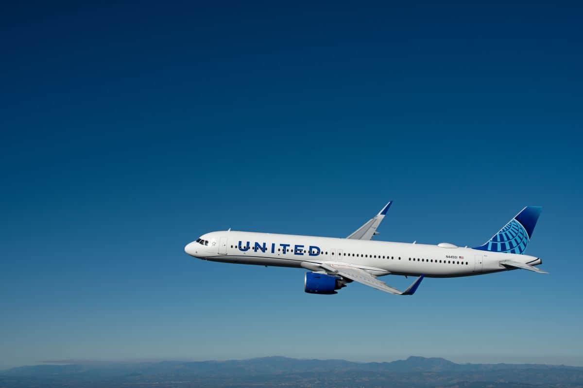 An aerial side-profile photograph of a united airlines boeing 737 max 9 jet in flight against a clear blue sky over a hazy mountainous landscape.