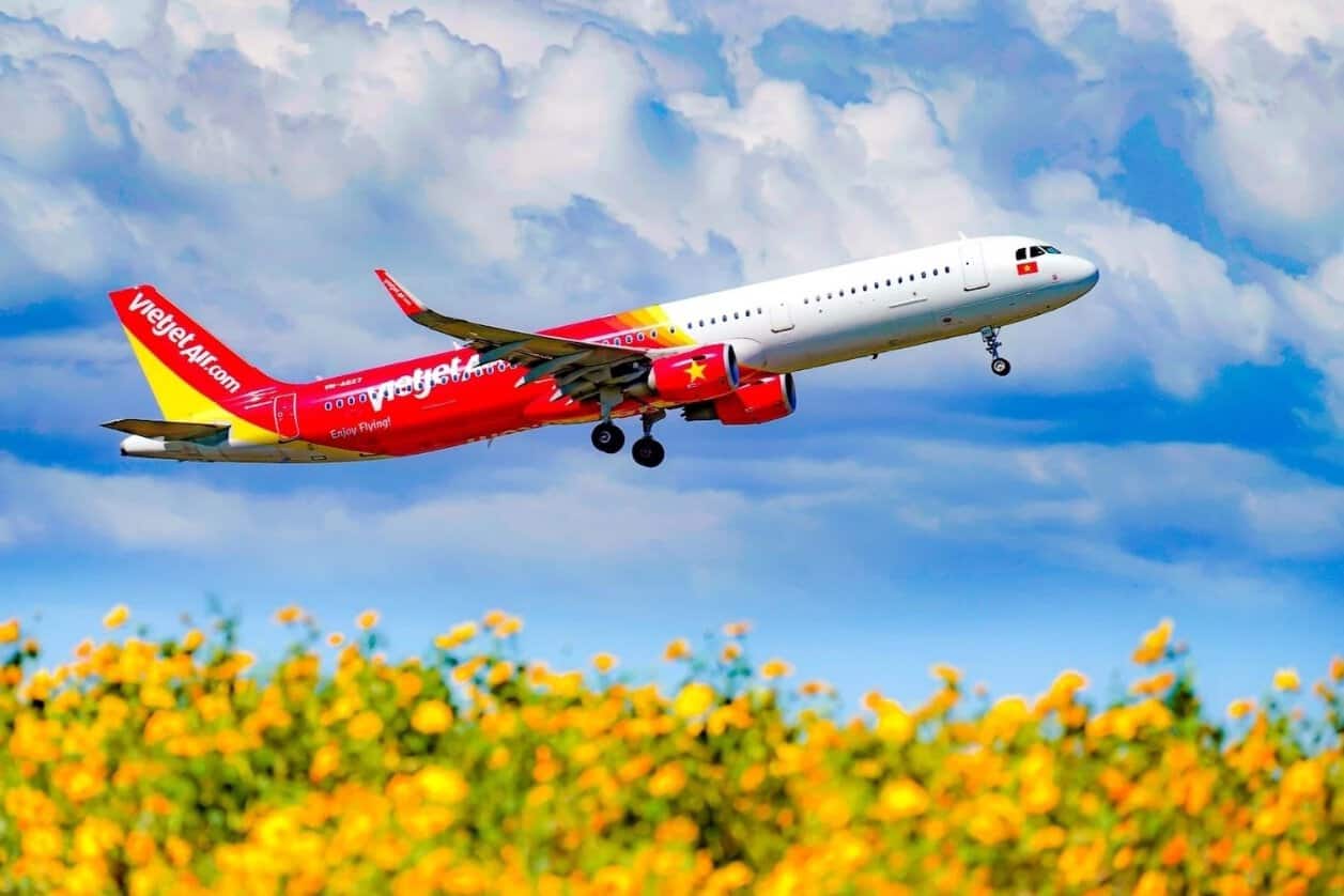 Vietjet aircraft with landing gear extended against a cloudy blue sky above yellow flowers.