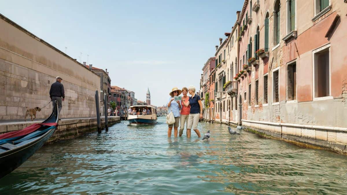 Venice visitor fee - travel and tour world Visitors check a travel itinerary beside a canal in venice.
