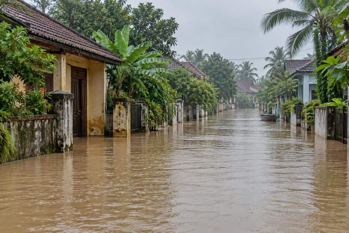 Vietnam faces travel disruption as local floods and landslide risks mean better route checks are now a must - travel and tour world Brown floodwater covers a vietnamese residential street lined with tiled-roof houses and tropical trees, with a small boat in the distance