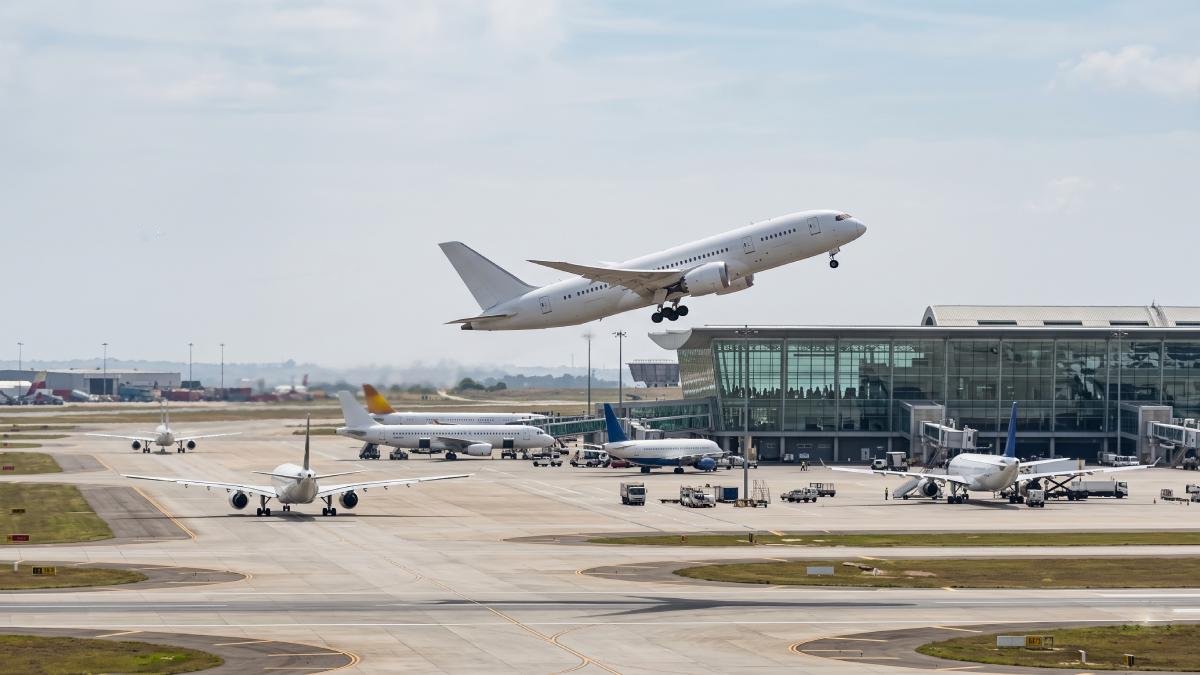 An editorial photograph of a modern white passenger jet taking off from a busy african international airport, with parked and taxiing aircraft, ground operations, and a contemporary glass terminal in the background.