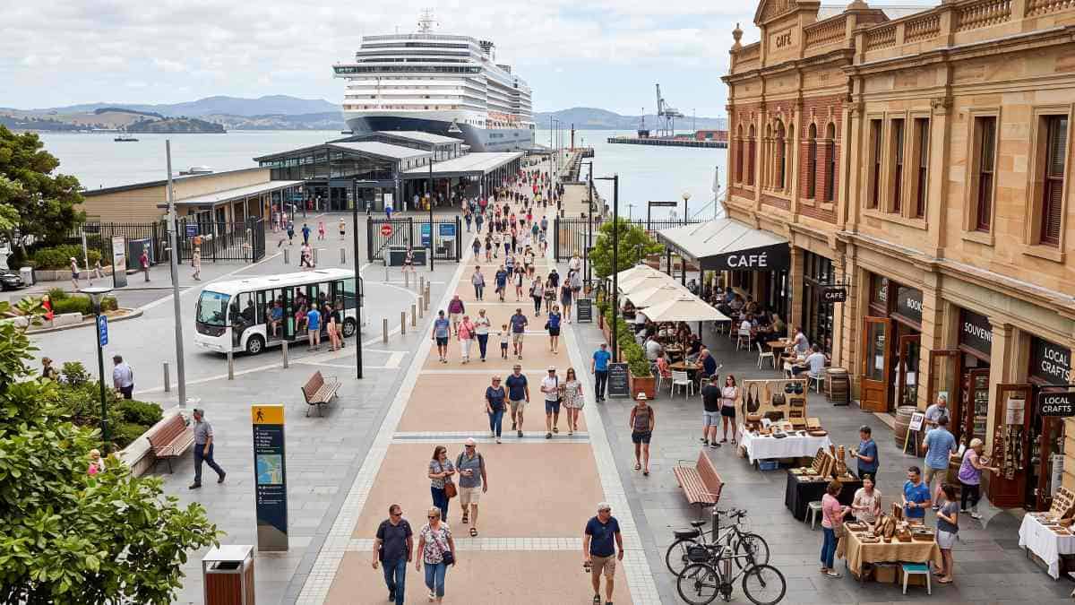 Cruise expenditure - travel and tour world Cruise passengers walk along a waterfront promenade from a docked ship toward outdoor cafés, local craft stalls, and a small electric shuttle.