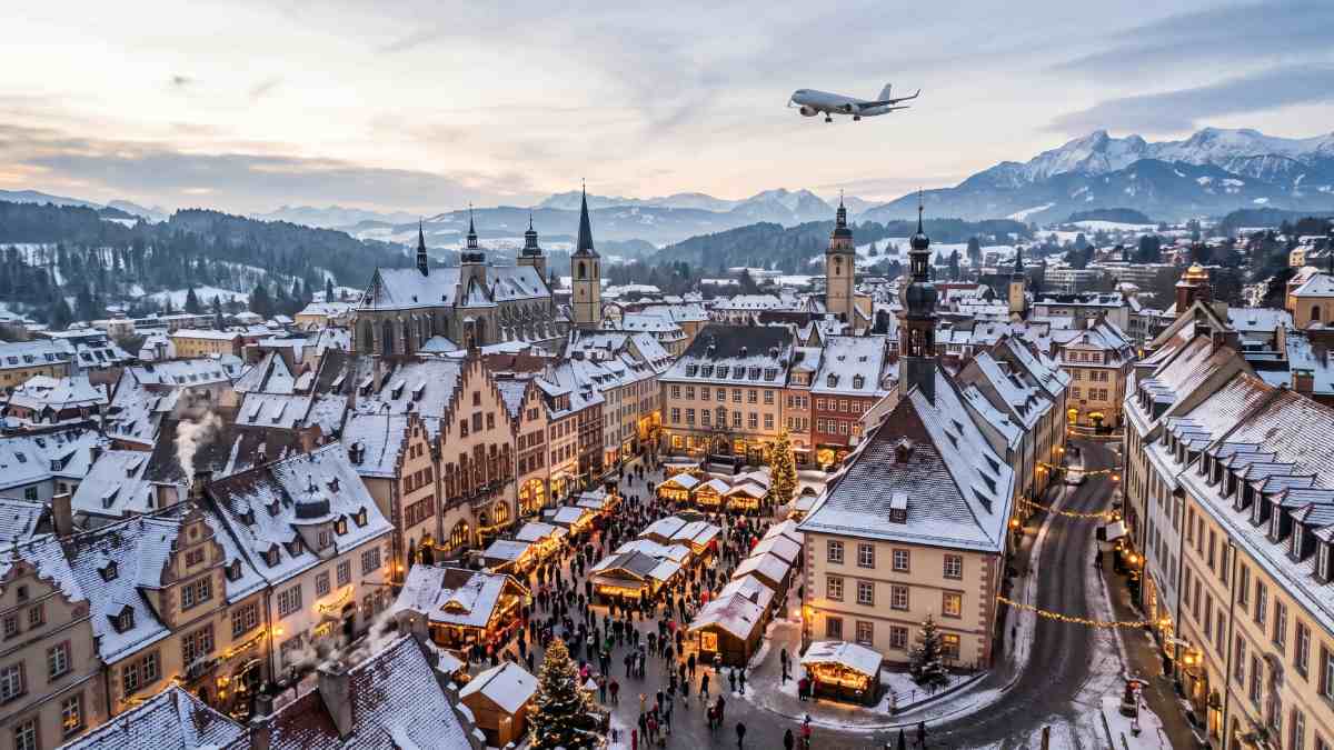 Flights expansions - travel and tour world A commercial passenger aircraft flies high over a snow-covered historic european city featuring a bustling, illuminated winter market and distant mountains.