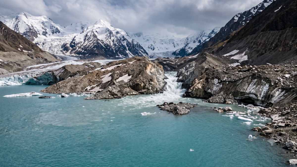 High-altitude glacial tourism - travel and tour world A small empty packraft floating on a turquoise glacial lake in the himalayas, with rushing meltwater cascading through rocky moraines and snow-capped peaks in the background.