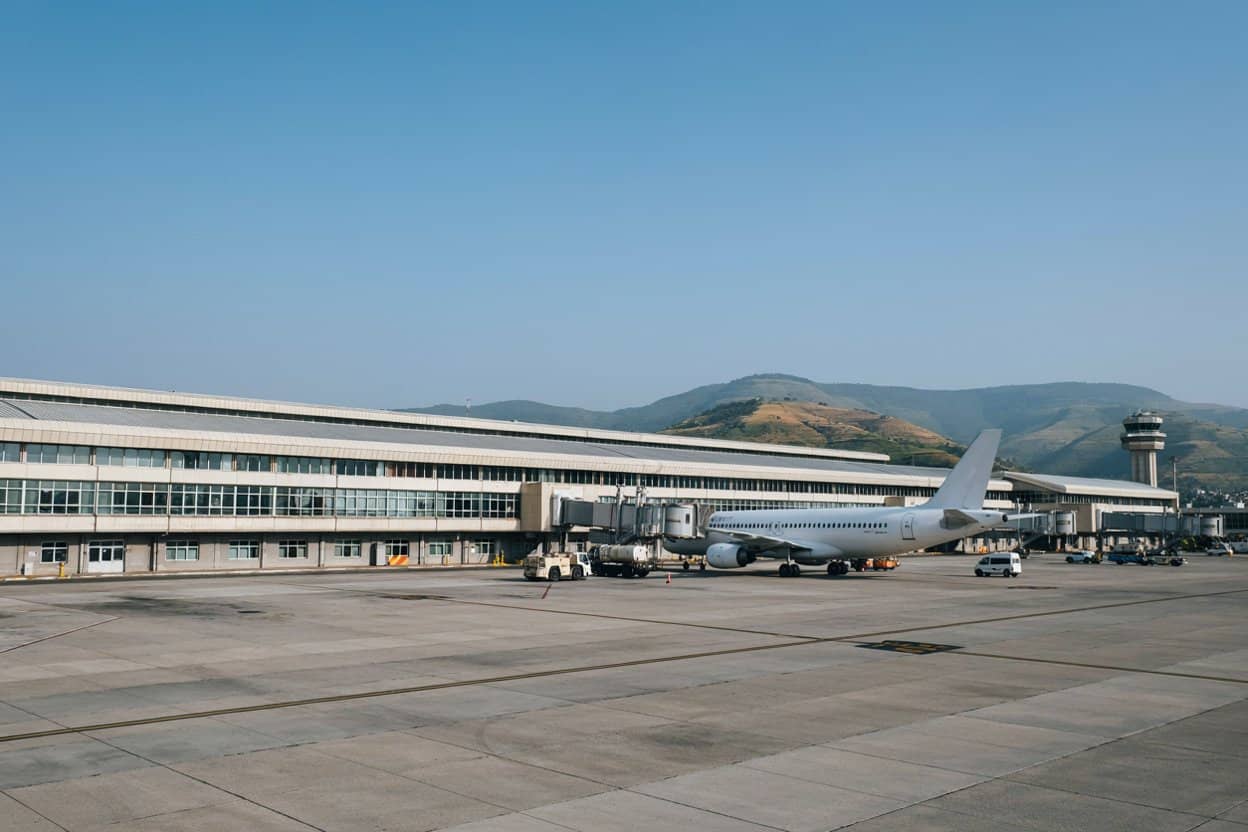 White passenger aircraft beside an airport terminal, with ground vehicles, a control tower and hills behind.