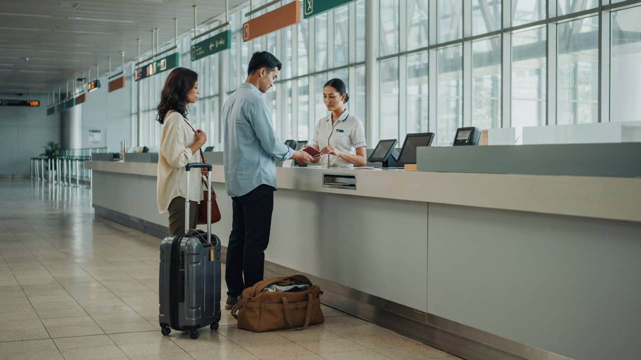Two travellers with luggage present travel documents to a staff member at an airport check-in counter