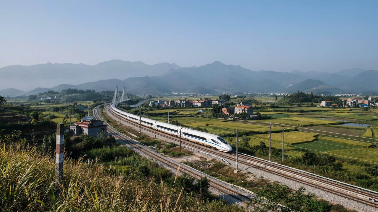 Rail travel - travel and tour world A sleek white bullet train moving along a curved track through rural farmland, with mountains stretching across the background