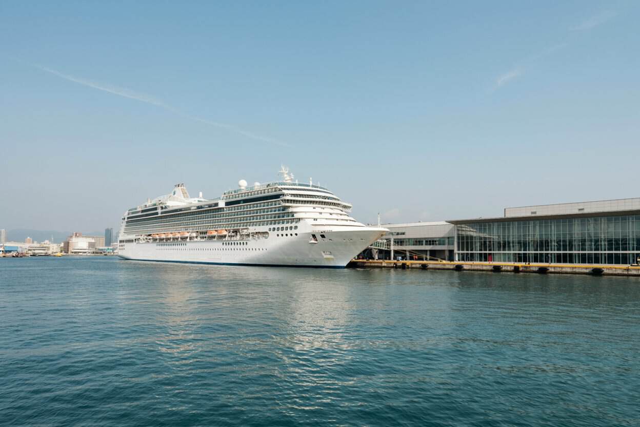 Japan is now using a new website to boost cruise tourism and reach one million japanese travellers by 2030 - travel and tour world Large white cruise ship docked beside a glass-fronted passenger terminal, with calm blue water and clear skies.