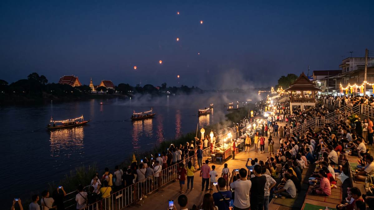 Crowds gather along an illuminated riverside promenade in thailand at night to watch reddish fireballs rise over the dark river, where traditional lit boats float past illuminated stalls and distant temples.