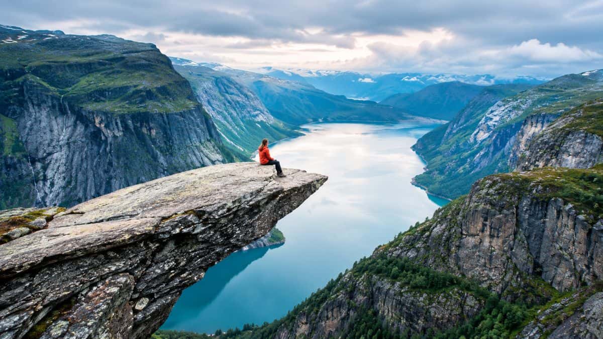 Hiker overlooking a dramatic fjord surrounded by rugged cliffs, green mountains, and misty peaks under a cloudy sky.