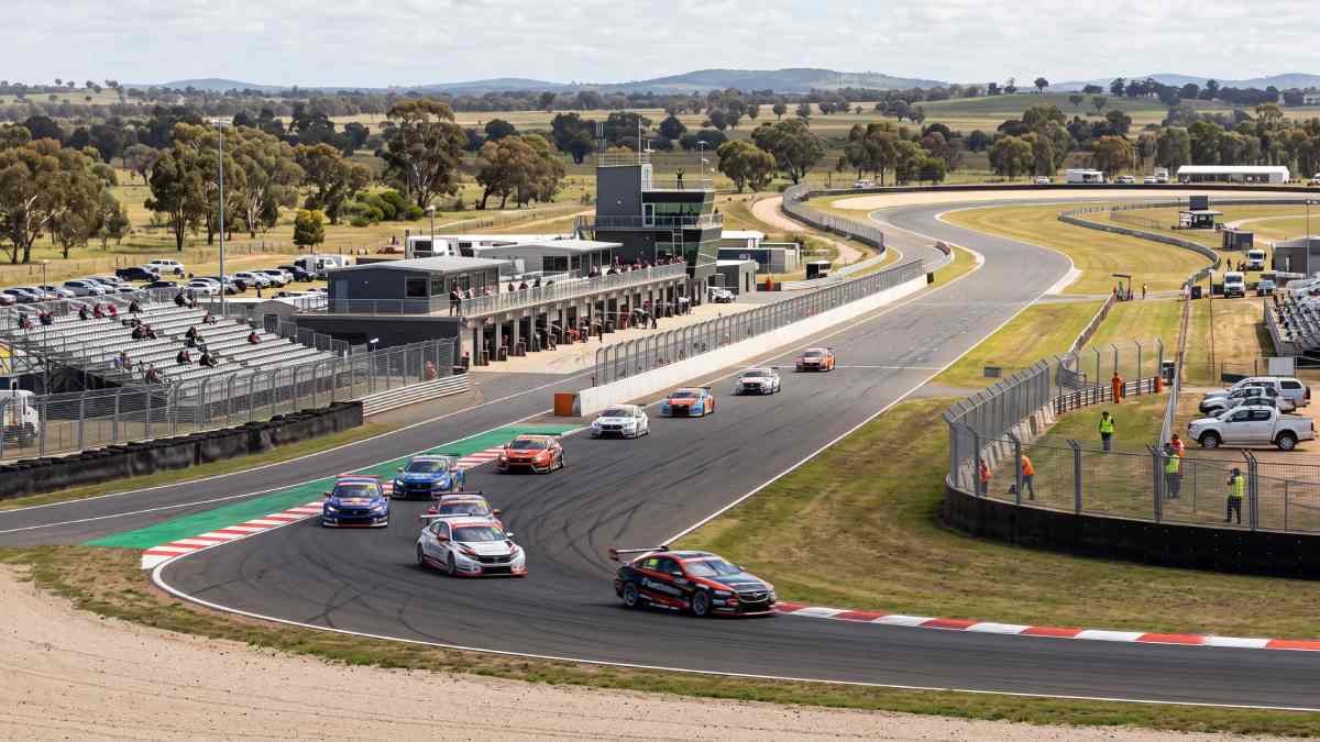 A fleet of touring racing cars corners along a sweeping paved circuit flanked by red-and-white kerbs, pit buildings, and rolling australian countryside under clear daylight.