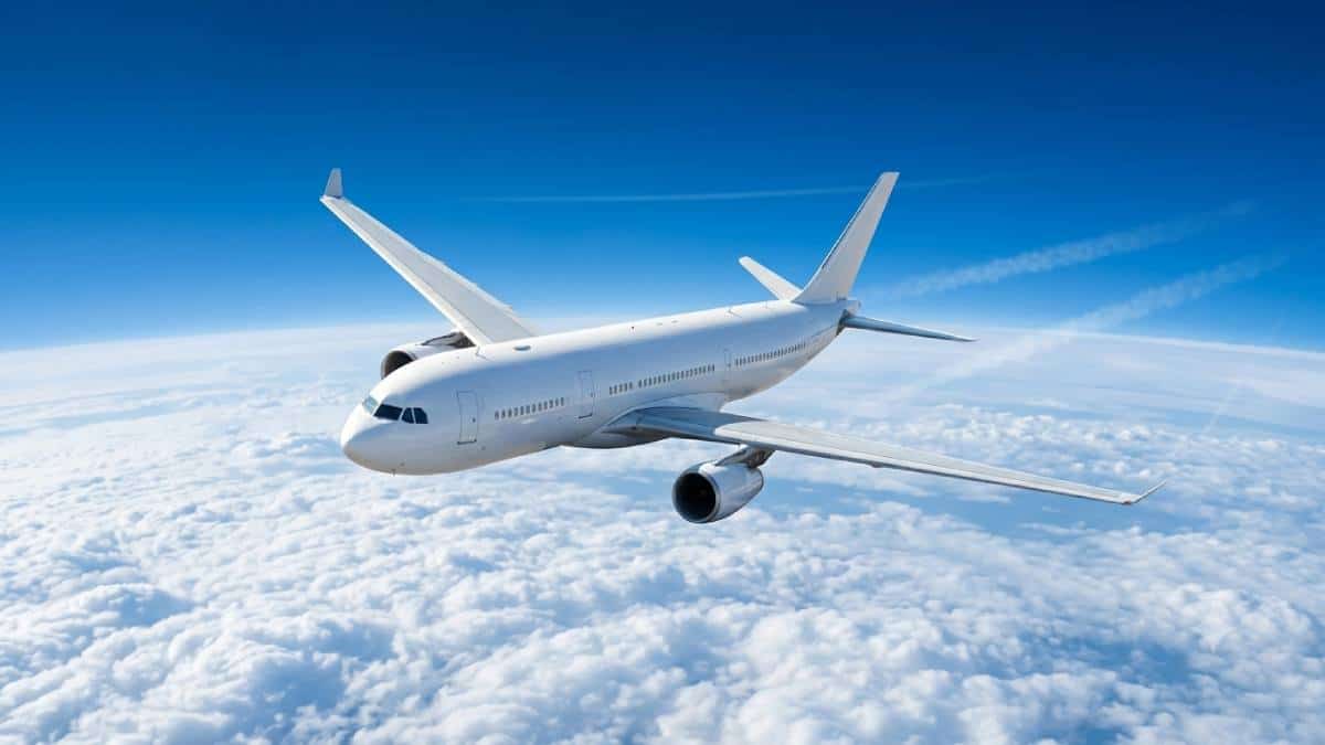 Tourism air routes - travel and tour world A white commercial passenger airplane flying high above a dense layer of fluffy white clouds against a clear blue sky, seen from a high three-quarter angle as it banks slightly during flight.